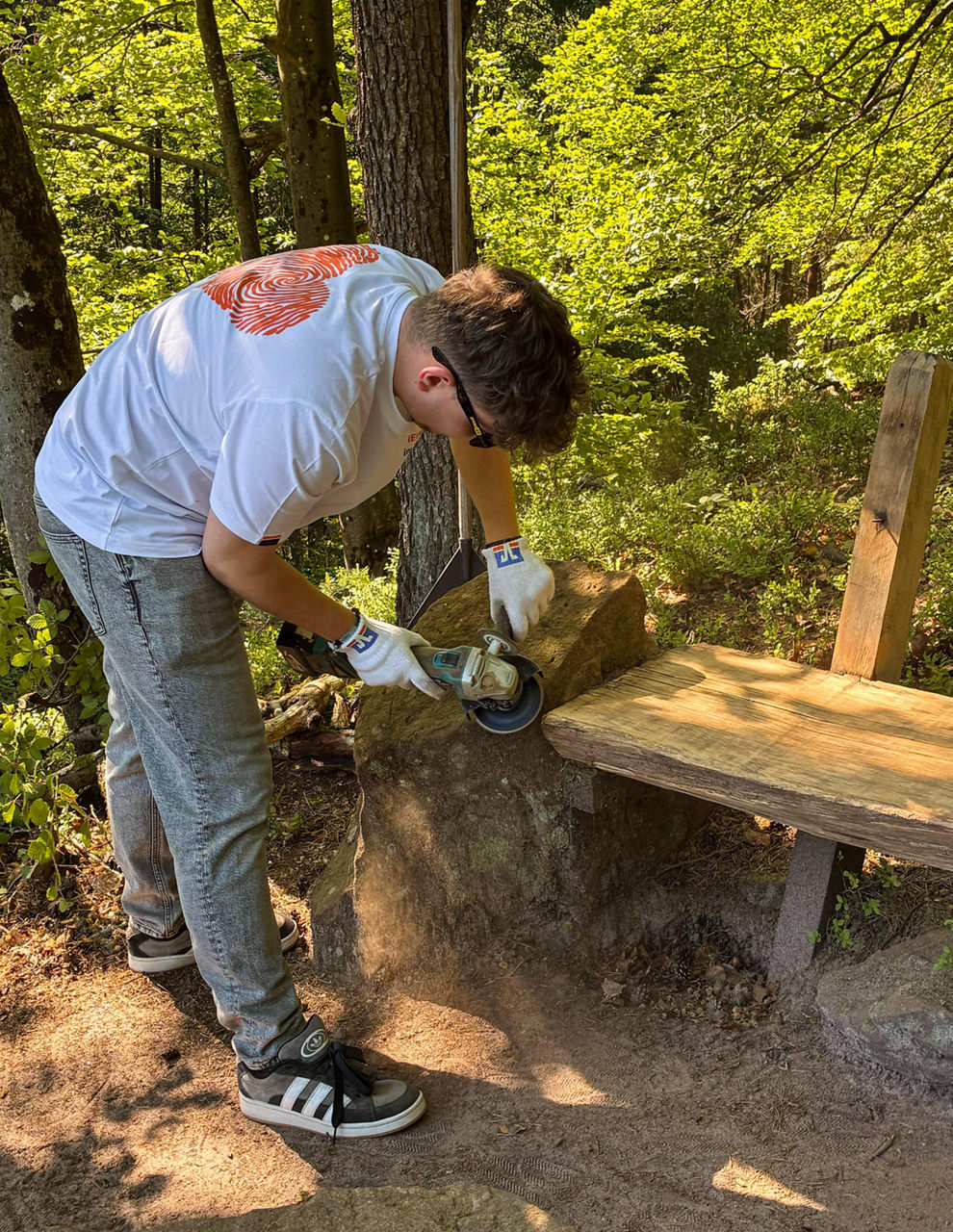 Eine Person arbeitet an einer Holzbank im Wald. Die Person nutzt ein Schleifgerät. Staub ist zu sehen.
