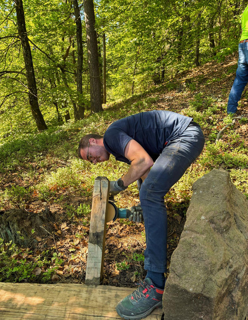 Eine Person arbeitet im Wald. Die Person schleift ein Stück Holz mit einer Maschine. Bäume sind rundherum.