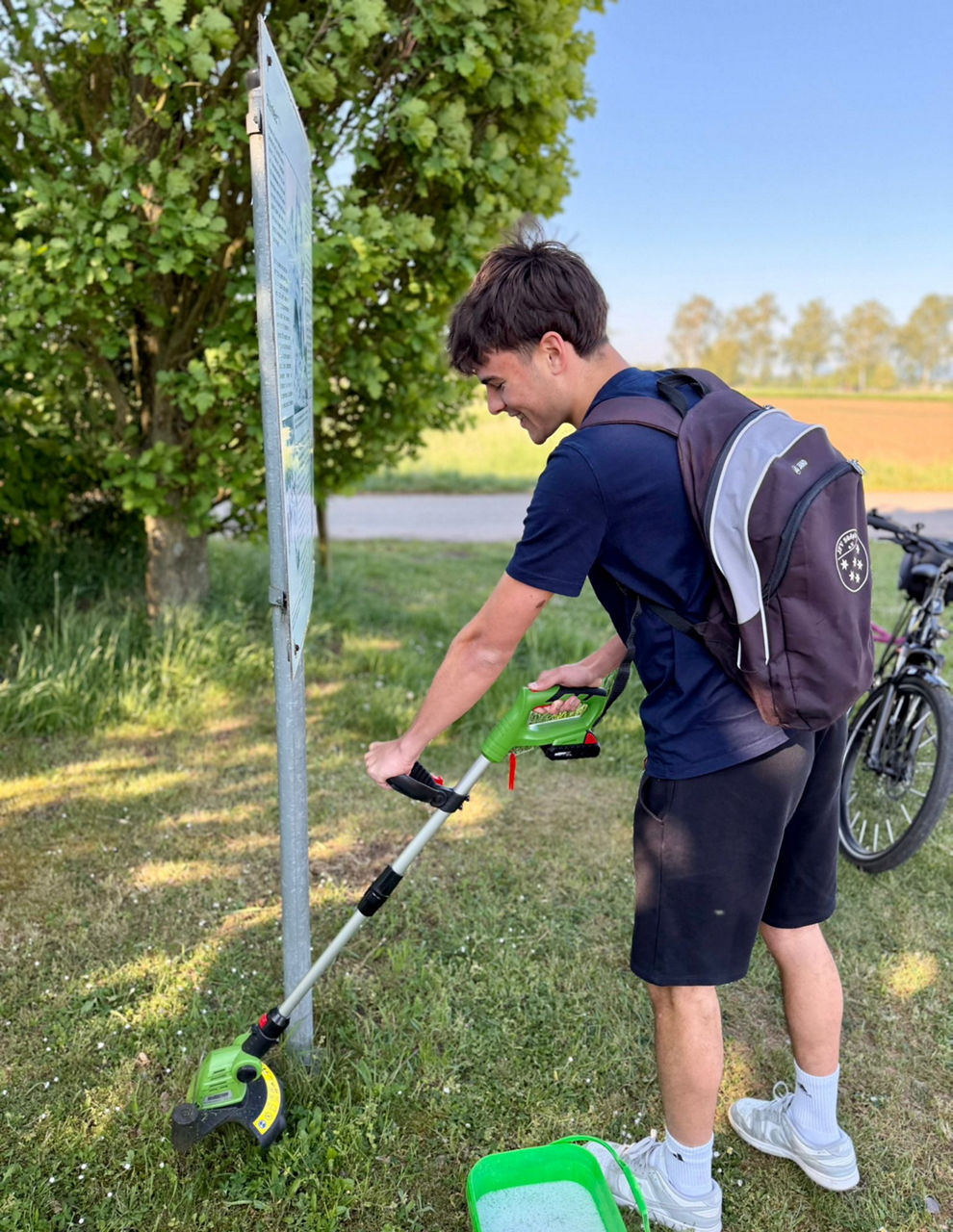 Person mit Rucksack nutzt einen Rasentrimmer und schneidet Gras neben einem Schild auf einer Wiese.