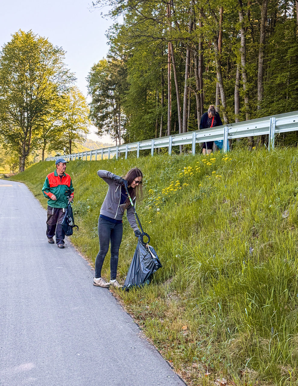 Drei Personen sammeln Abfall an einer Böschung neben einer Straße mit Greifzangen und Müllsäcken.