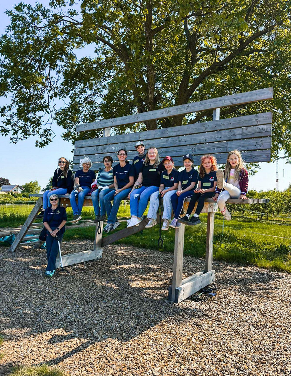 Mehrere Personen sitzen zusammen auf einer großen Holzbank draußen. Es gibt Gras und Bäume im Hintergrund.