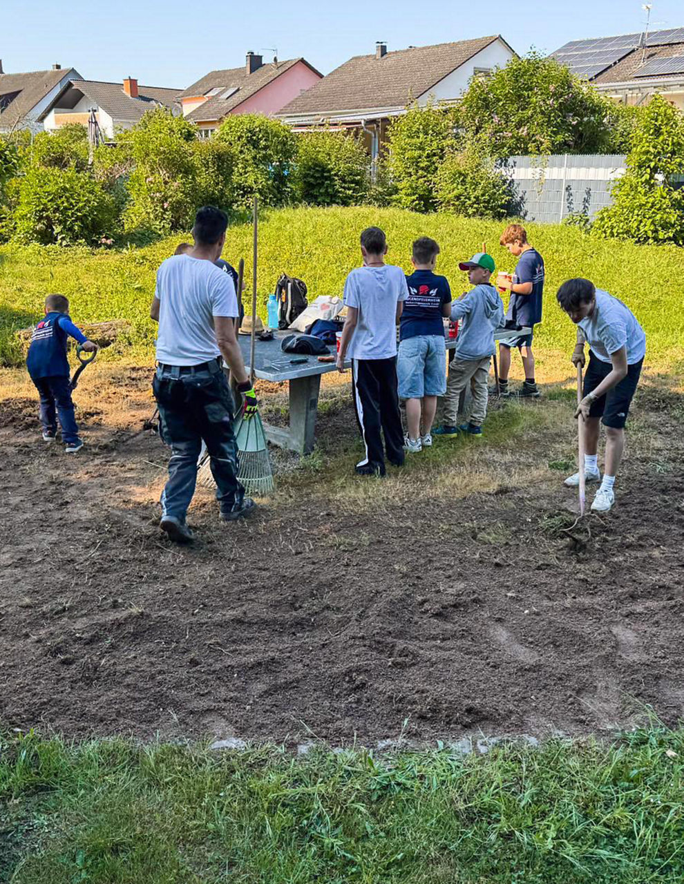 Mehrere Menschen arbeiten draußen auf einer Fläche mit Erde. Sie nutzen Rechen und Schaufeln. Ein Tisch steht dabei.