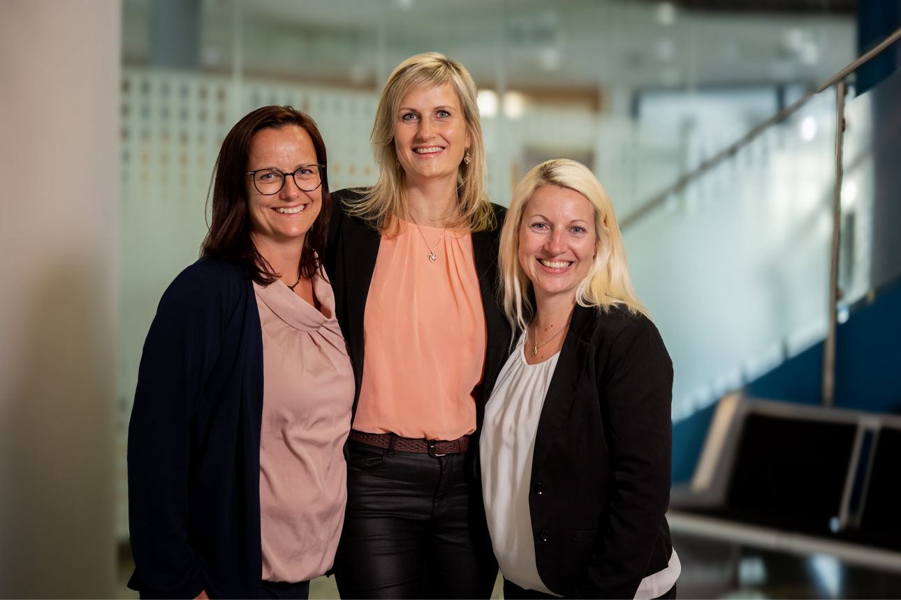 3 Frauen stehen in einem Schalterraum und lächeln