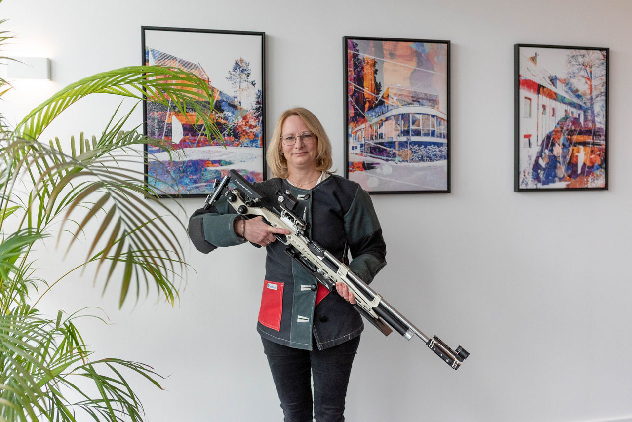 Frau steht mit Schießjacke und Luftwehr in einem Raum mit einer Blume und drei Bildern an der Wand
