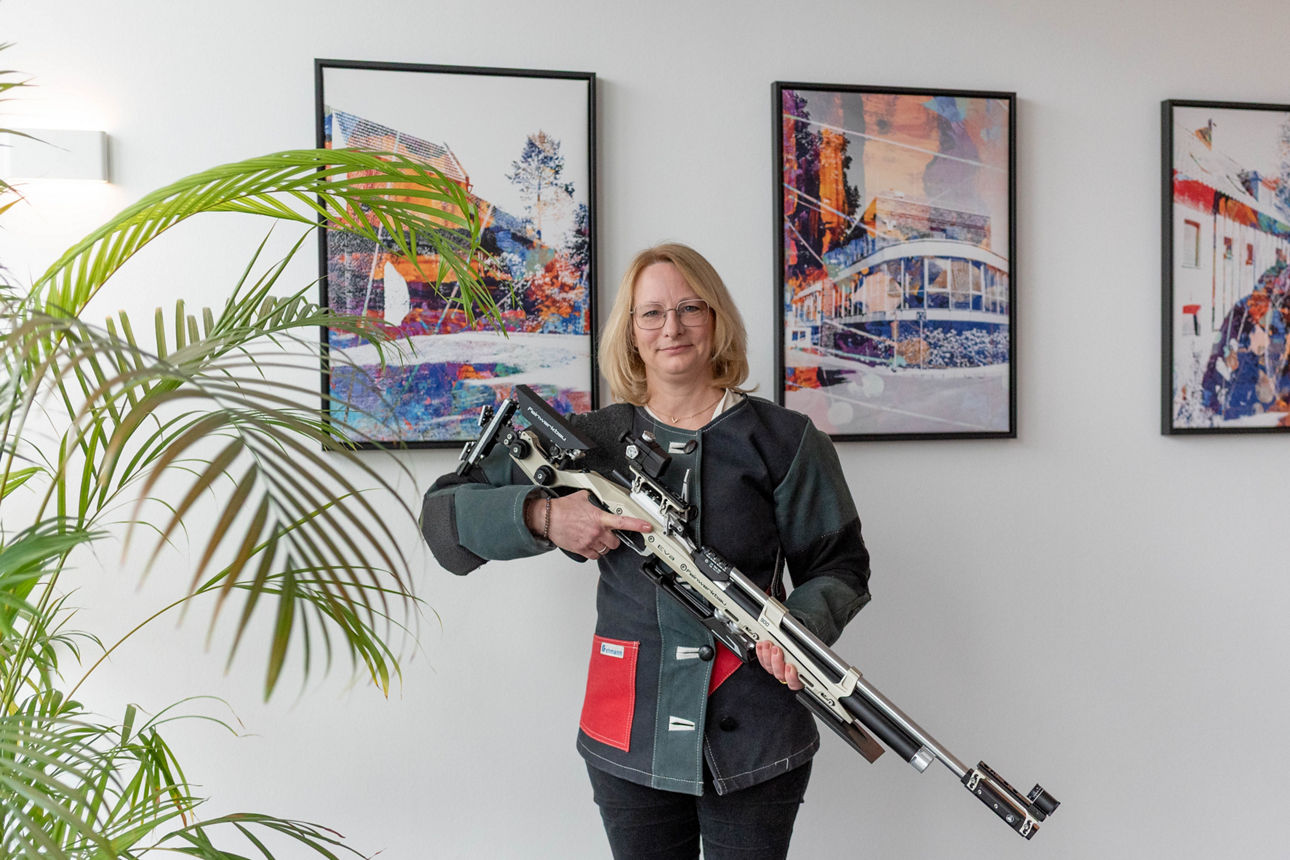 Frau steht mit Schießjacke und Luftwehr in einem Raum mit einer Blume und drei Bildern an der Wand