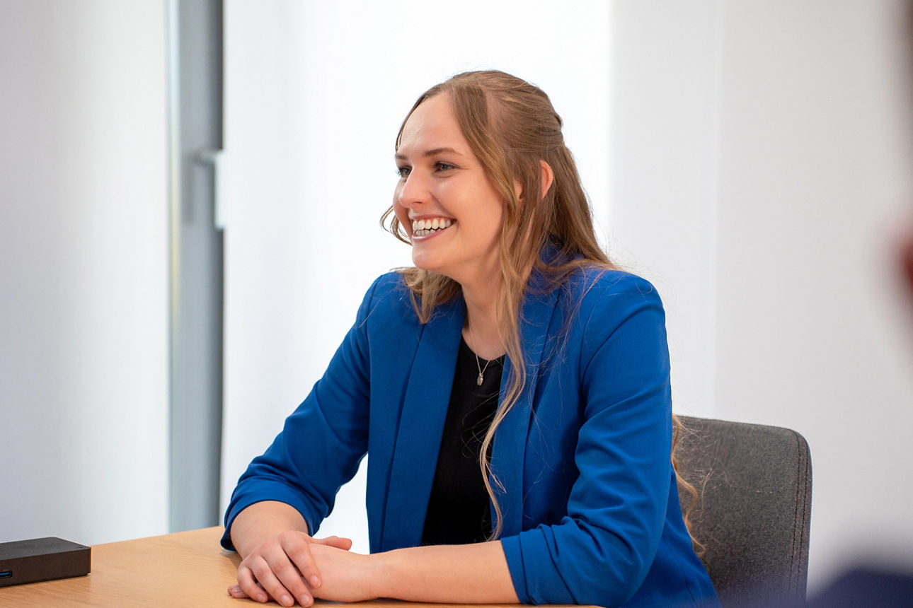Eine blonde, junge Frau mit blauem Blazer sitzt an einem Tisch und  lächelt ihr Gegenüber an, welches nicht mehr im Bild zu sehen ist. Die Szene spielt in einem Beratungszimmer der Volksbank Ruhr Mitte.