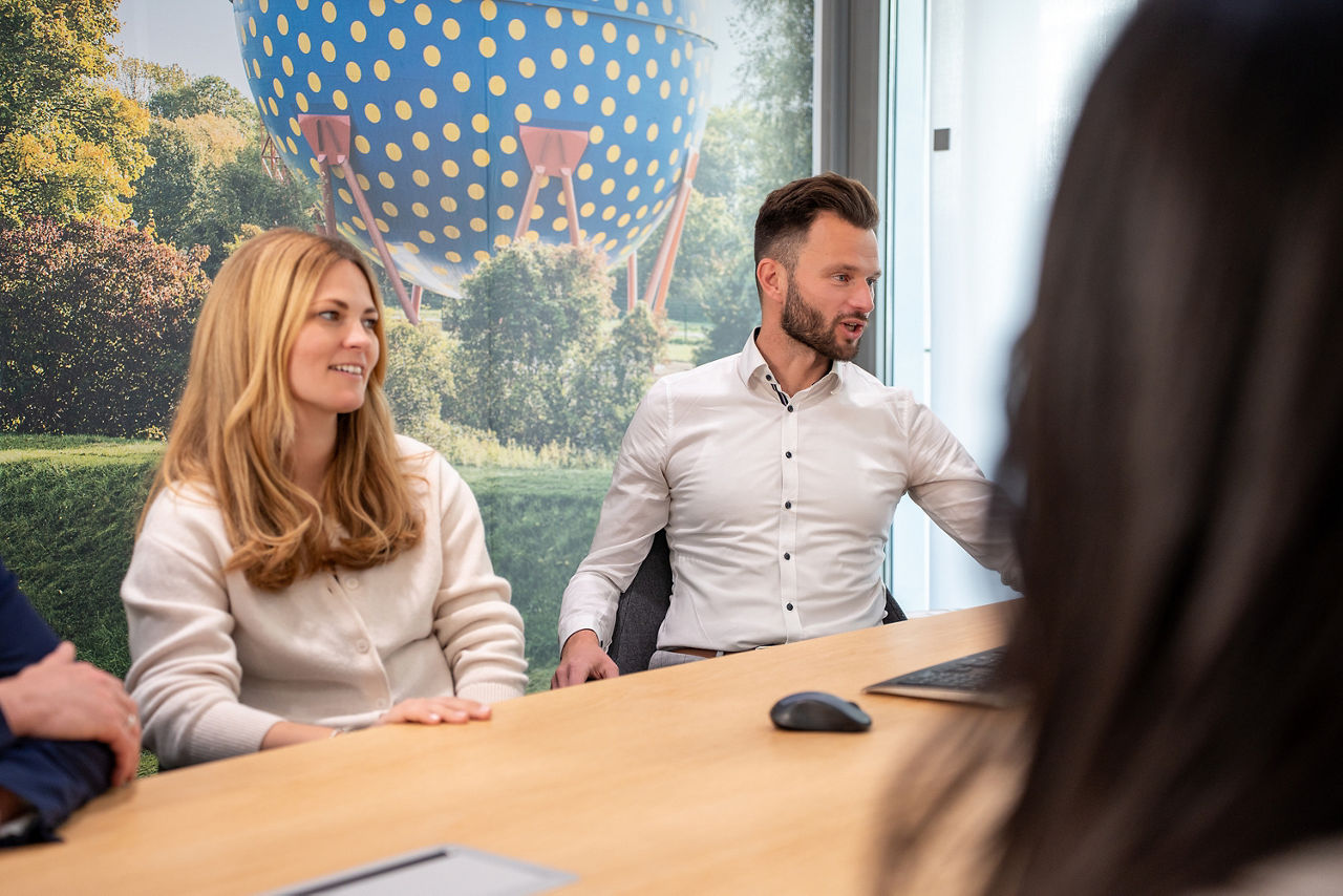 Eine junge, blonde Frau sitzt an einem Tisch in einem modernen Beratungszimmer und schaut freundlich auf einen Monitor außerhalb des Bildes. Links neben ihr sitzt angedeutet eine männliche Person, ihr gegenüber und nur von hinten angedeutet eine weitere weibliche Person. Rechts neben ihr sitzt ein freundlicher junger Kundenberater in weißem Hemd mit Bart, welcher Ebenfalls auf den Monitor außerhalb des Bildes schaut.
