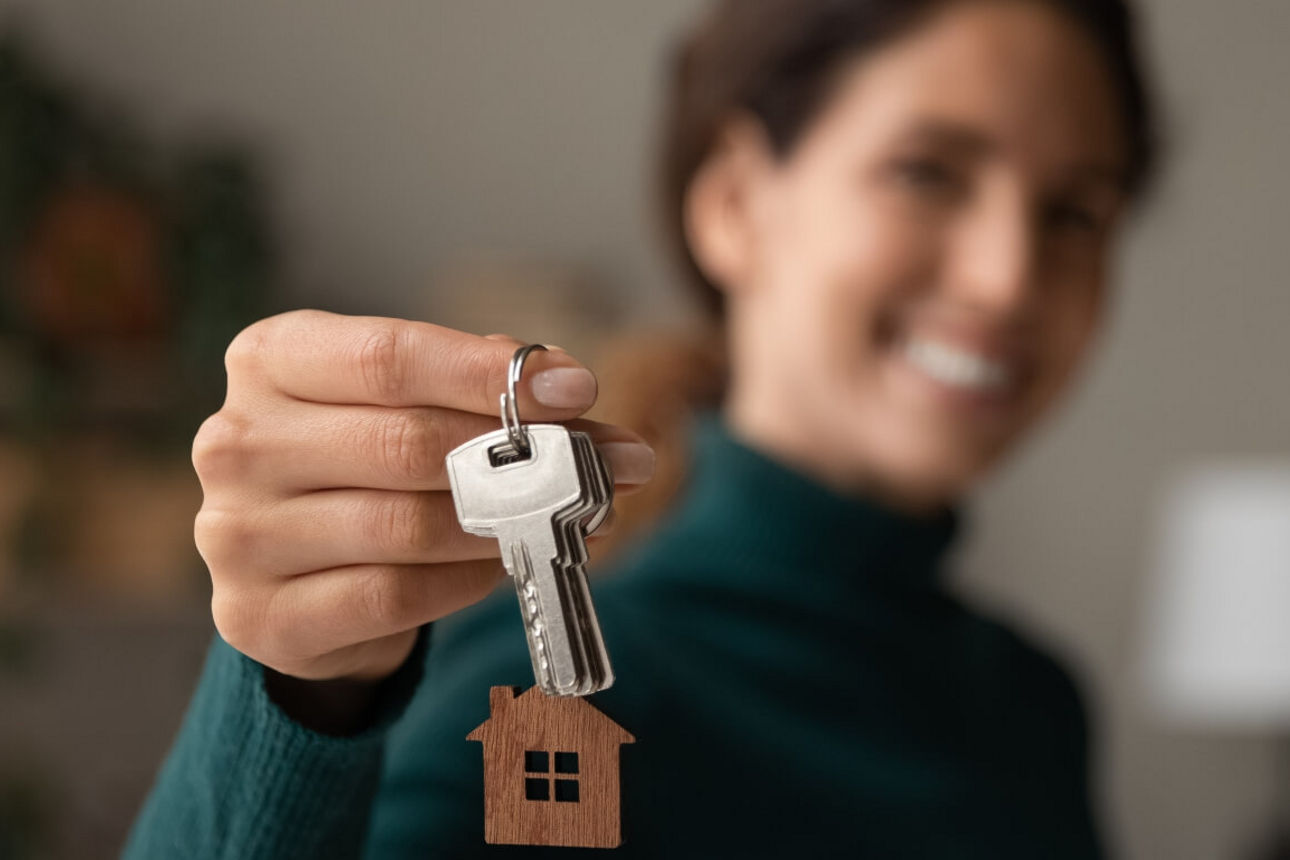 Frau hält Schlüsselbund mit Schlüsselanhänger in Hausform in die Kamera. Frau im Hintergrund ist unscharf