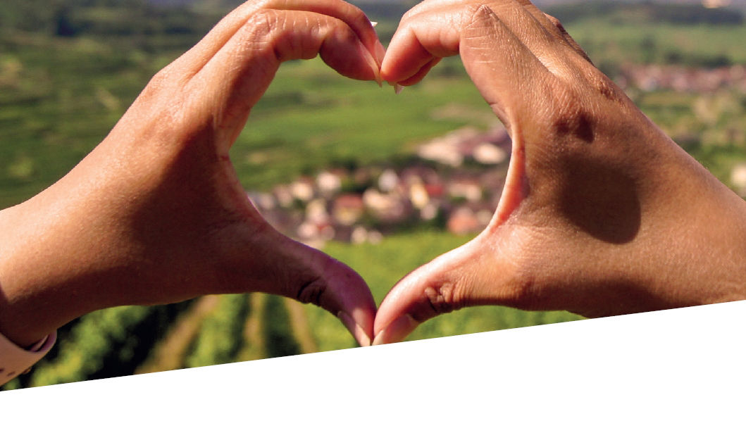 Zwei Hände formen ein Herz vor einer grünen Hügellandschaft – Symbol für Verbundenheit und Liebe zur Region.