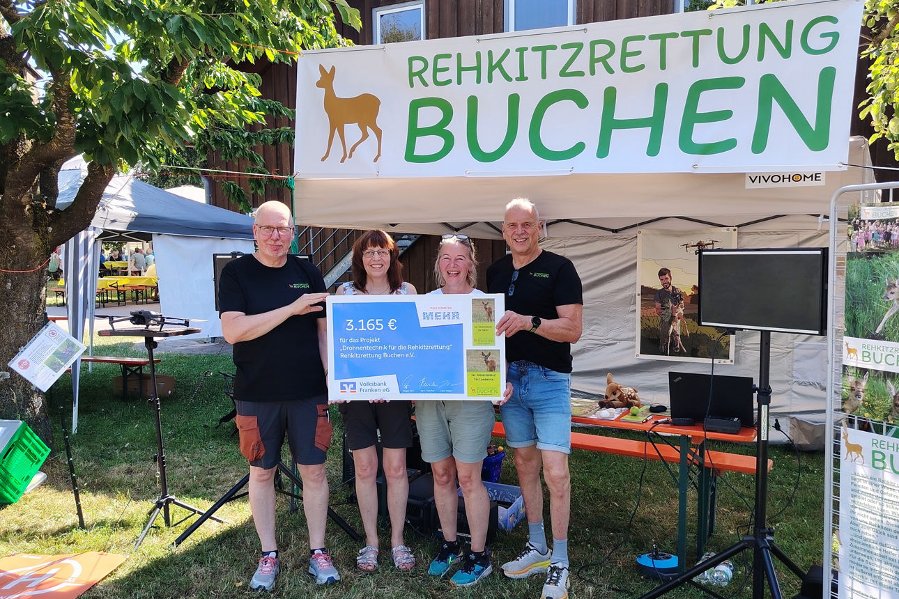 ier Personen stehen lächelnd vor dem Stand „Rehkitzrettung Buchen“ bei einem Outdoor‑Fest und halten gemeinsam einen großen Spendenscheck über 3.165 € für Drohnentechnik hoch.