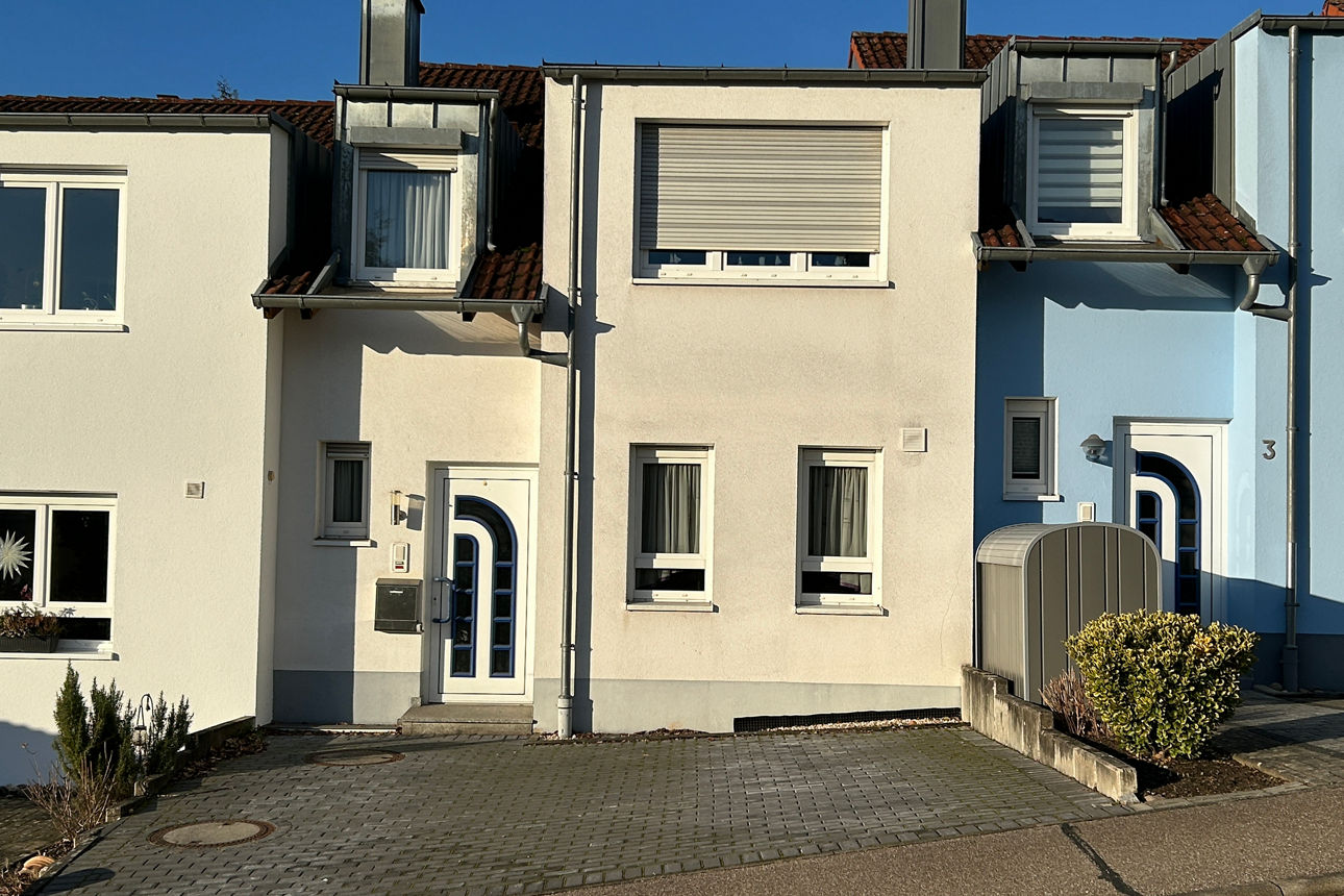 Reihenhäuser mit hellen Fassaden, Haustüren und Rollläden an einer ruhigen Wohnstraße bei Tageslicht.