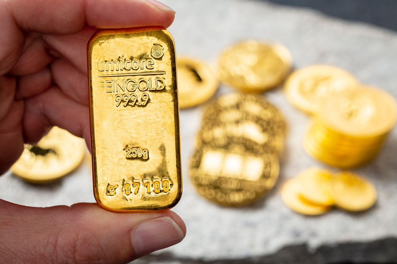 Eine Hand hält einen Goldbarren in der Hand. Im Hintergrund sind Goldmünzen zu sehen