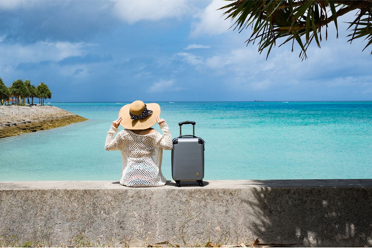 Frau mit Hut und Koffer sitzt auf einer Mauer an einem paradiesischen Strand