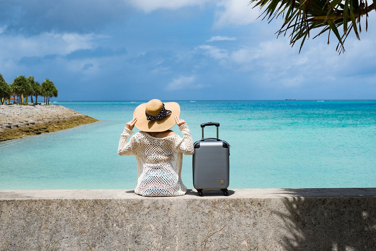Frau mit Hut und Koffer sitzt auf einer Mauer an einem paradiesischen Strand