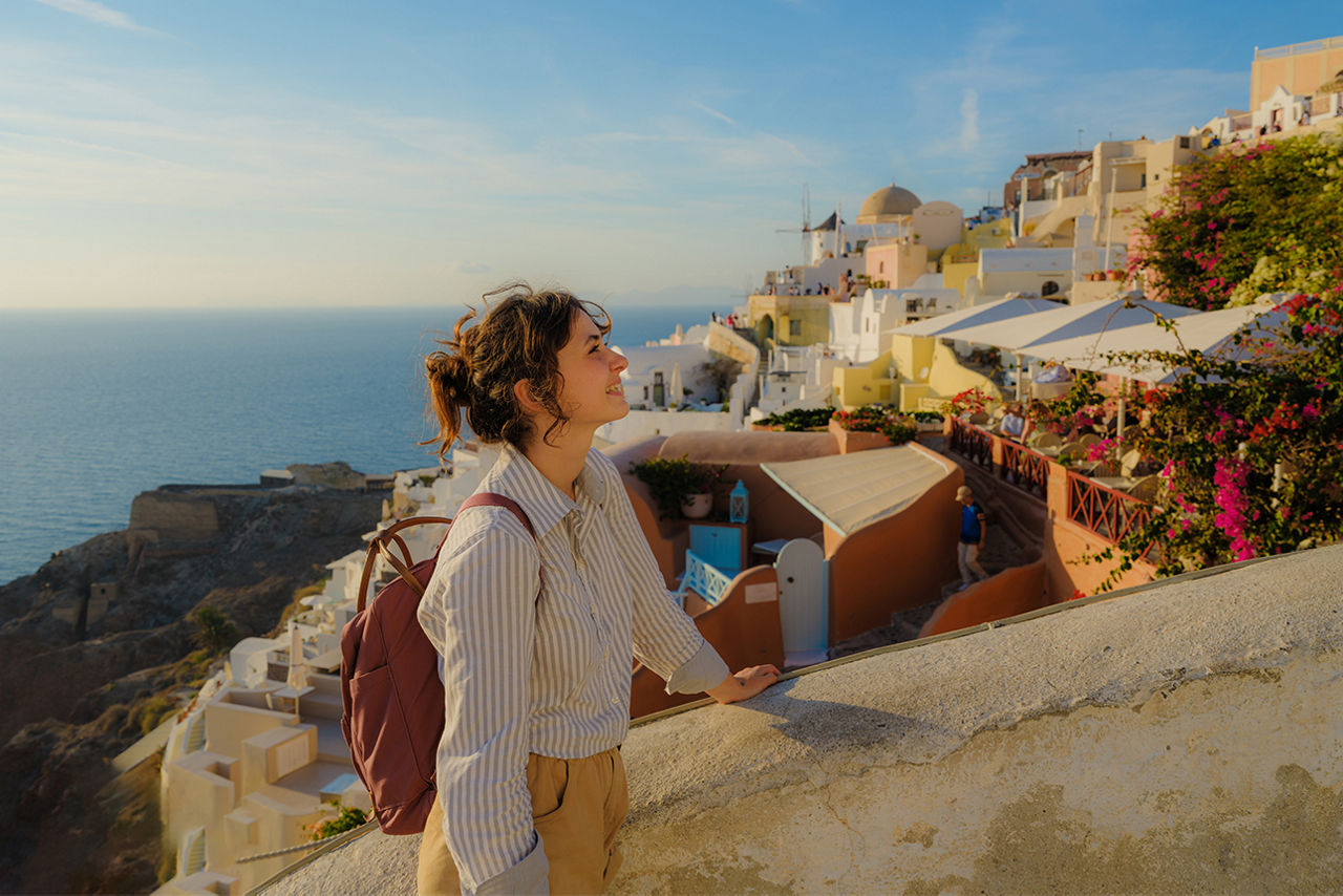 Frau im Urlaub lehnt sich an einer Mauer an. Im Hintegrund sieht man  Häuser und das Meer.