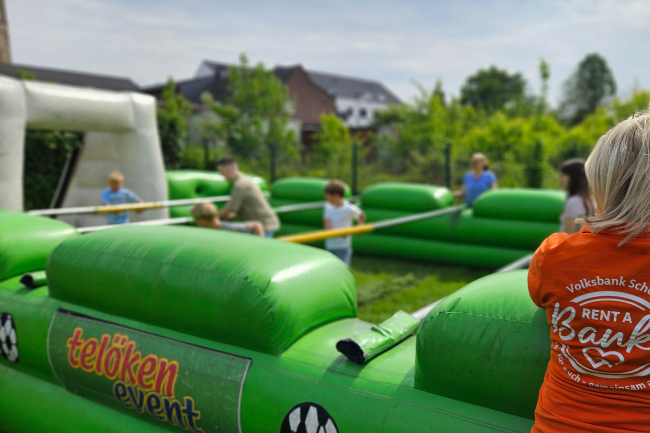Ein Mitglied des Rent a Banker Teams lehnt an einem aufblasbaren, grünen Menschenkicker. Einige Jugendliche spielen dort.