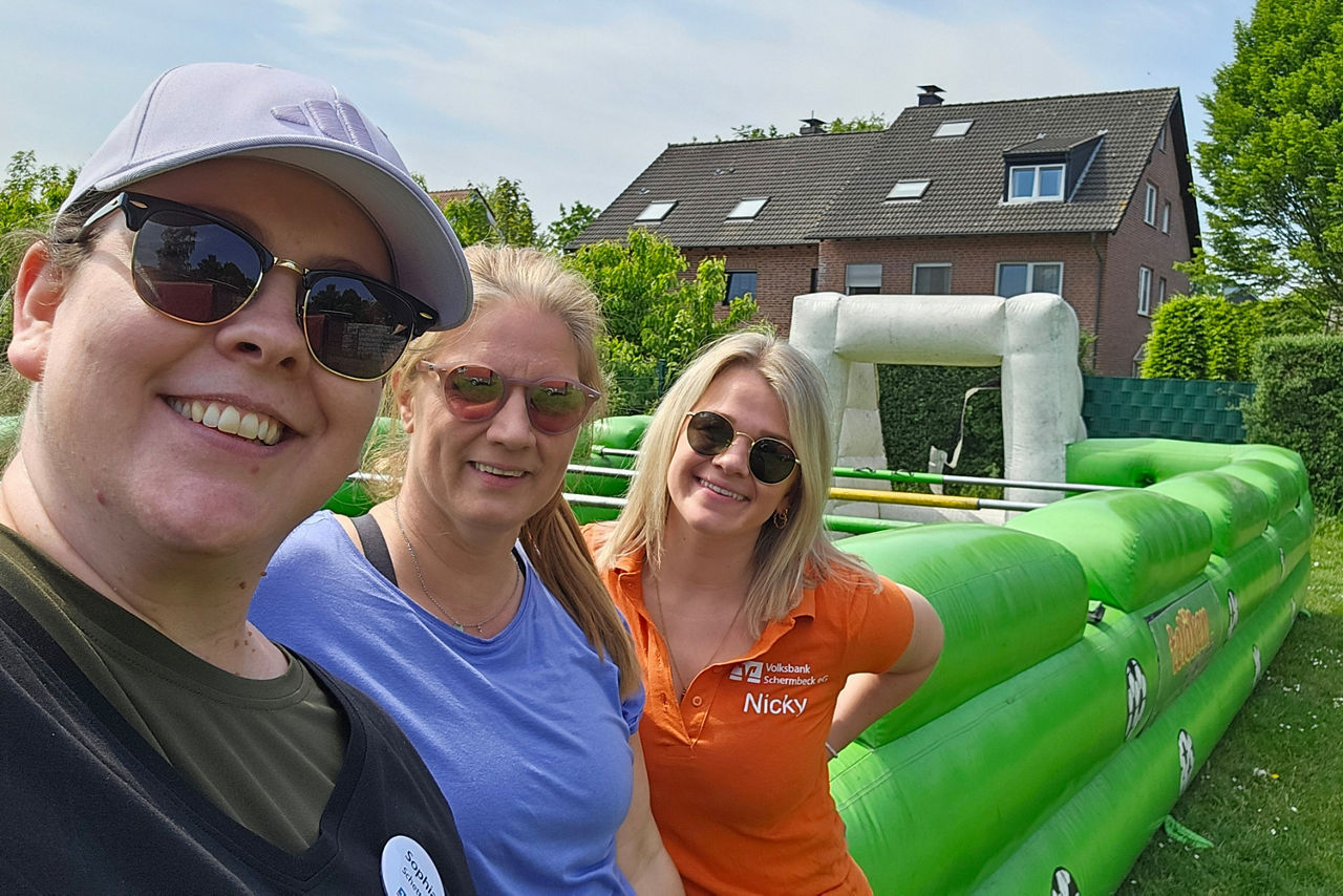 Drei weibliche Mitglieder des Rent a Banker Teams vor dem aufblasbaren, grünen Menschenkicker.