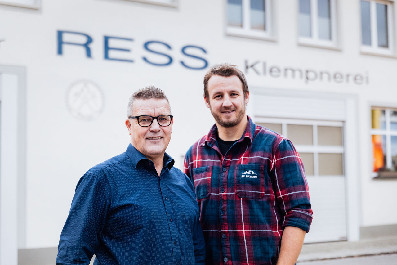 Georg Rützel und Christoph Rützel stehen vor dem Firmengebäude des Sanitär‑ und Klempnerbetriebs der „Ress GmbH“. Sichtbar ist die Gebäudebeschriftung des Unternehmens sowie der Außenbereich des Betriebs