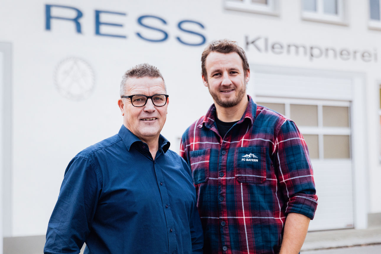 Georg Rützel und Christoph Rützel stehen vor dem Firmengebäude des Sanitär‑ und Klempnerbetriebs der „Ress GmbH“. Sichtbar ist die Gebäudebeschriftung des Unternehmens sowie der Außenbereich des Betriebs