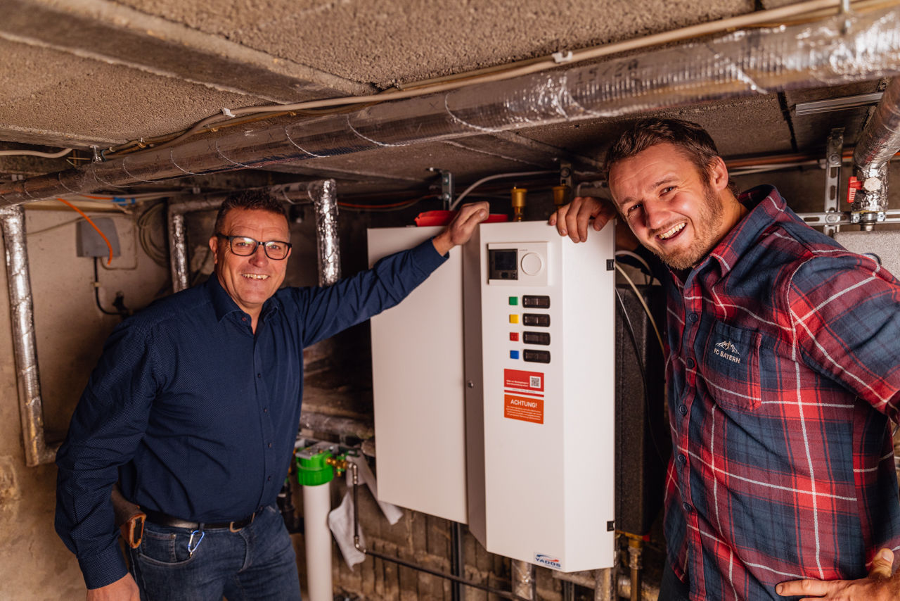 Georg Rützel und Christoph Rützel stehen im Technikraum des Sanitär‑ und Heizungsbetriebs von „Ress GmbH“. Sichtbar sind montierte Heizungs‑ und Regeltechnik, Rohrleitungen sowie verschiedene Installationsbauteile aus dem Bereich der Gebäudetechnik
