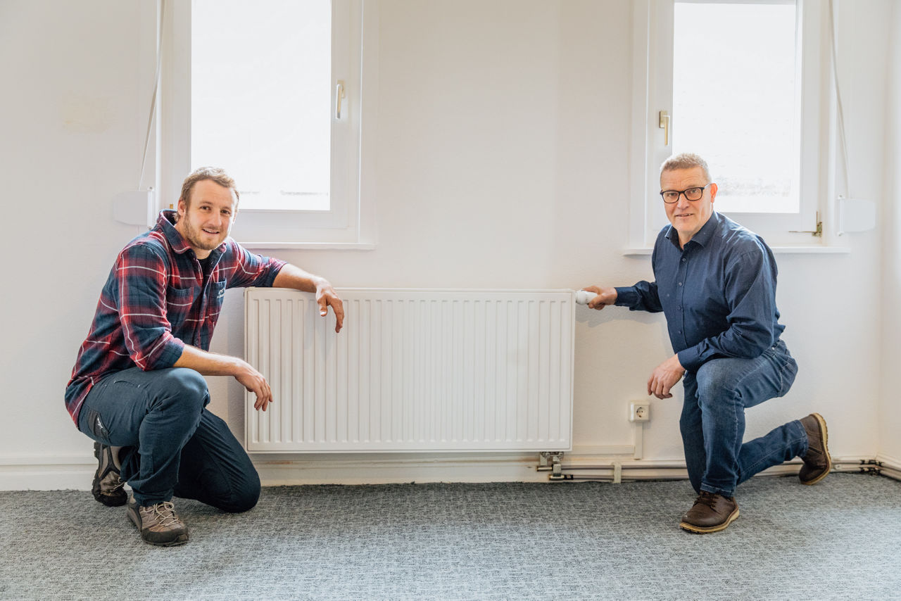 Christoph Rützel und Georg Rützel stehen vor einem montierten Heizkörper im Sanitär‑ und Gebäudetechnikbereich des Unternehmens „Ress GmbH“. Sichtbar sind zudem Rohrleitungen und weitere installierte Komponenten der Heizungsanlage