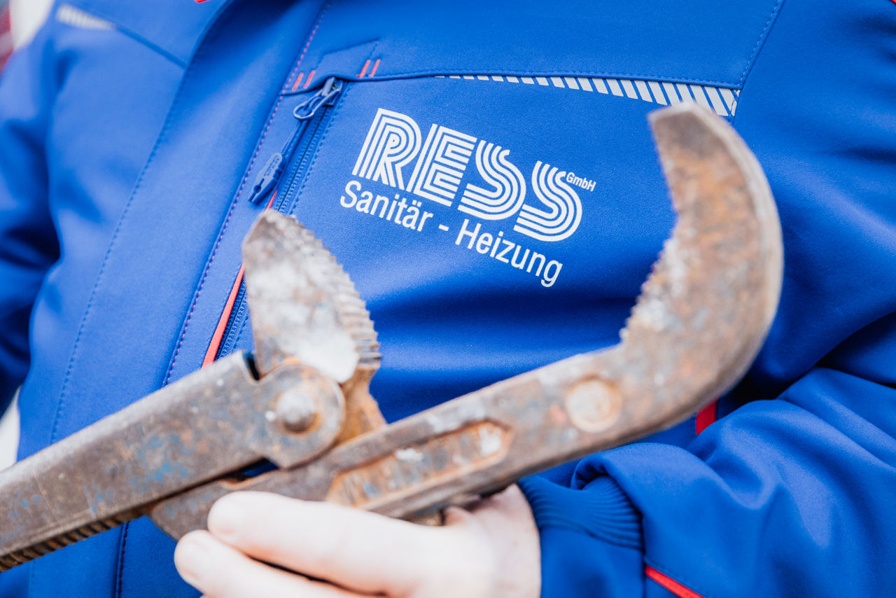 Christoph Rützel und Georg Rützel stehen vor einem montierten Heizkörper im Sanitär‑ und Gebäudetechnikbereich des Unternehmens „Ress GmbH“. Sichtbar sind zudem Rohrleitungen und weitere installierte Komponenten der Heizungsanlage