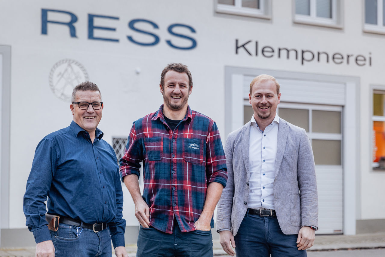 Georg Rützel, Christoph Rützel und Berater Florian Abschütz stehen vor dem Firmengebäude des Sanitär‑ und Heizungsbetriebs der „Ress GmbH“. Sichtbar sind die Gebäudebeschriftung sowie der Eingangsbereich des Betriebs
