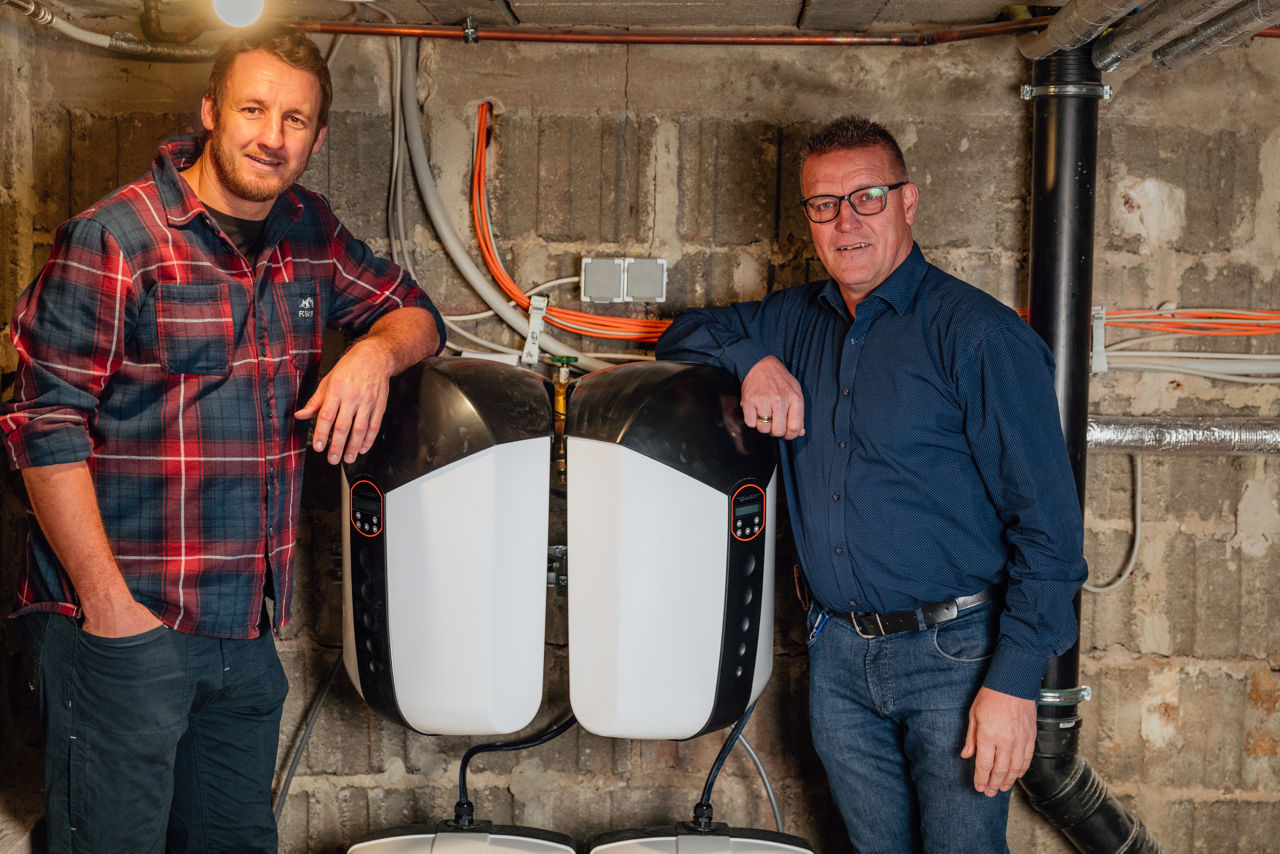Christoph Rützel und Georg Rützel stehen im Technikraum des Sanitär‑ und Heizungsbetriebs der „Ress GmbH“. Zu sehen sind zwei nebeneinander montierte Wärmetechnik‑Module, umgeben von sichtbaren Leitungen und Installationen aus dem Bereich des Sanitär‑ und Heizungsbaus