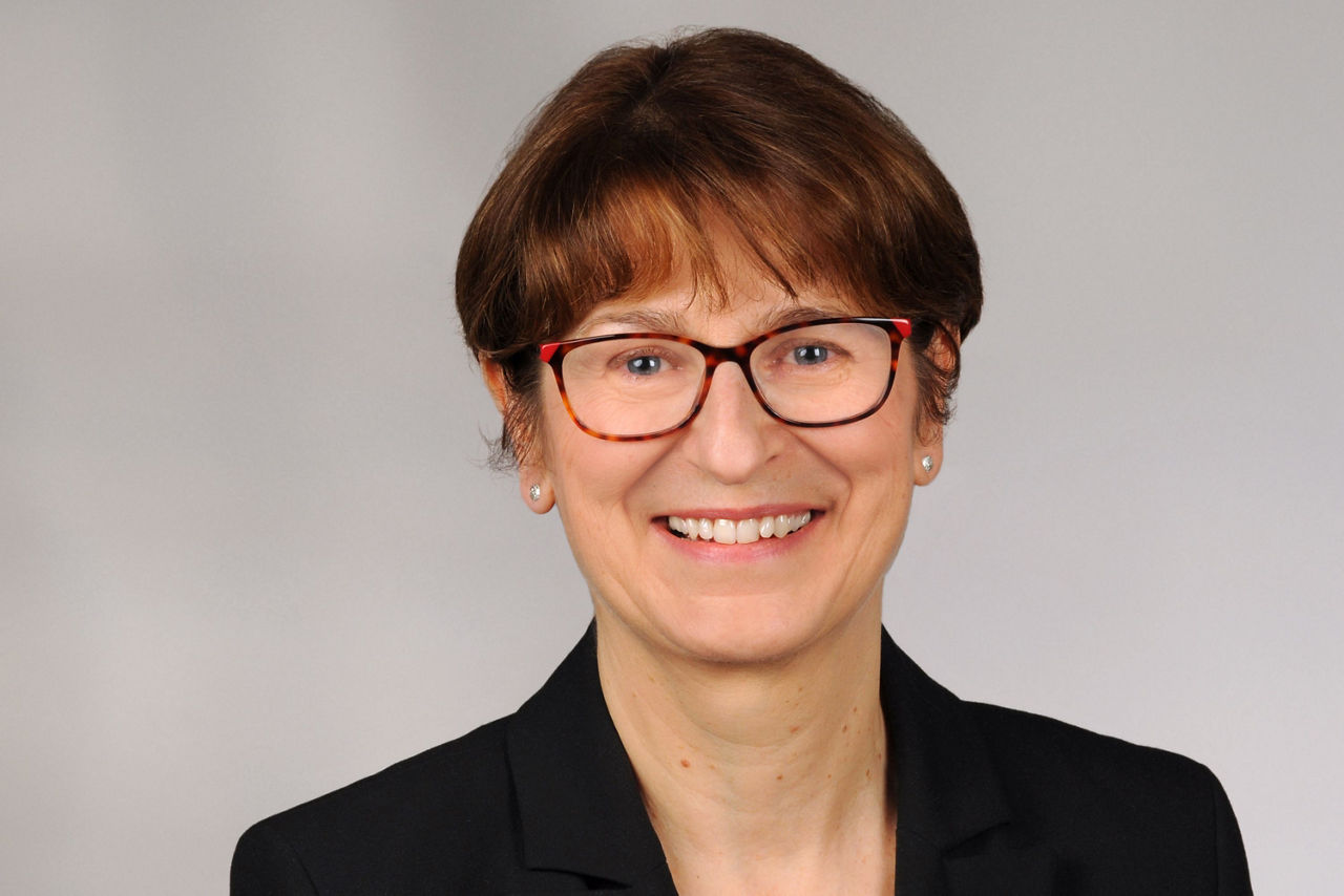 Frau mit kurzen braunen Haaren, trägt schwarzen Blazer sowie eine Brille und Perlenohrringe, vor neutralem Hintergrund