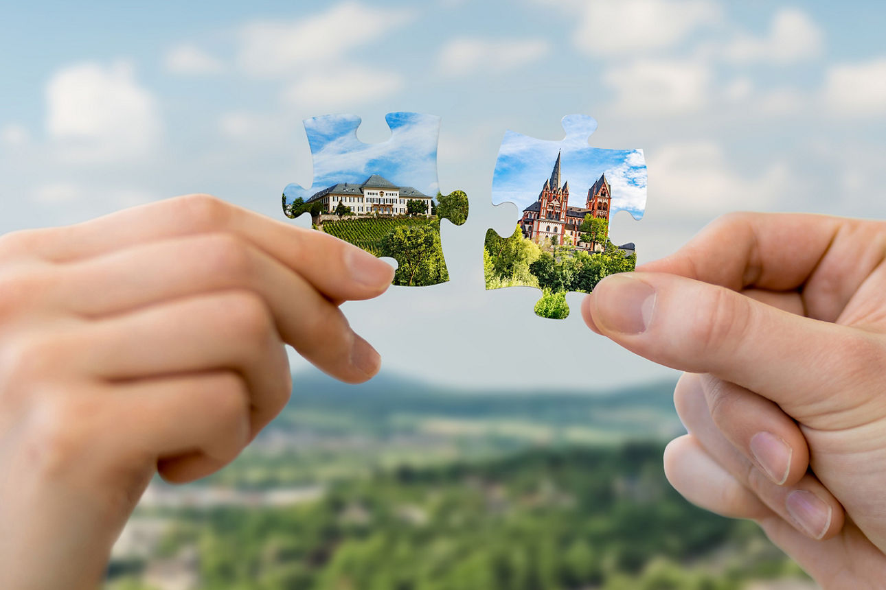 Zwei Hände halten jeweils ein Puzzlestück mit Bildern von Gebäuden und Landschaften vor unscharfem Himmel