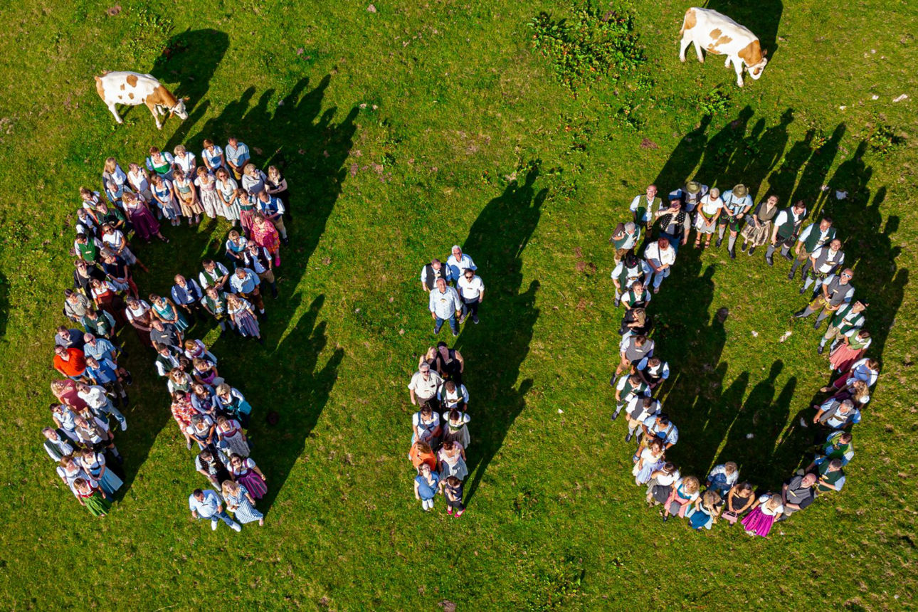 Eine Gruppe von Menschen in Trachtenkleidung bildet auf einer grünen Wiese die Buchstaben "R I O". Drei Kühe grasen in der Nähe. Die Aufnahme ist aus der Vogelperspektive gemacht.
