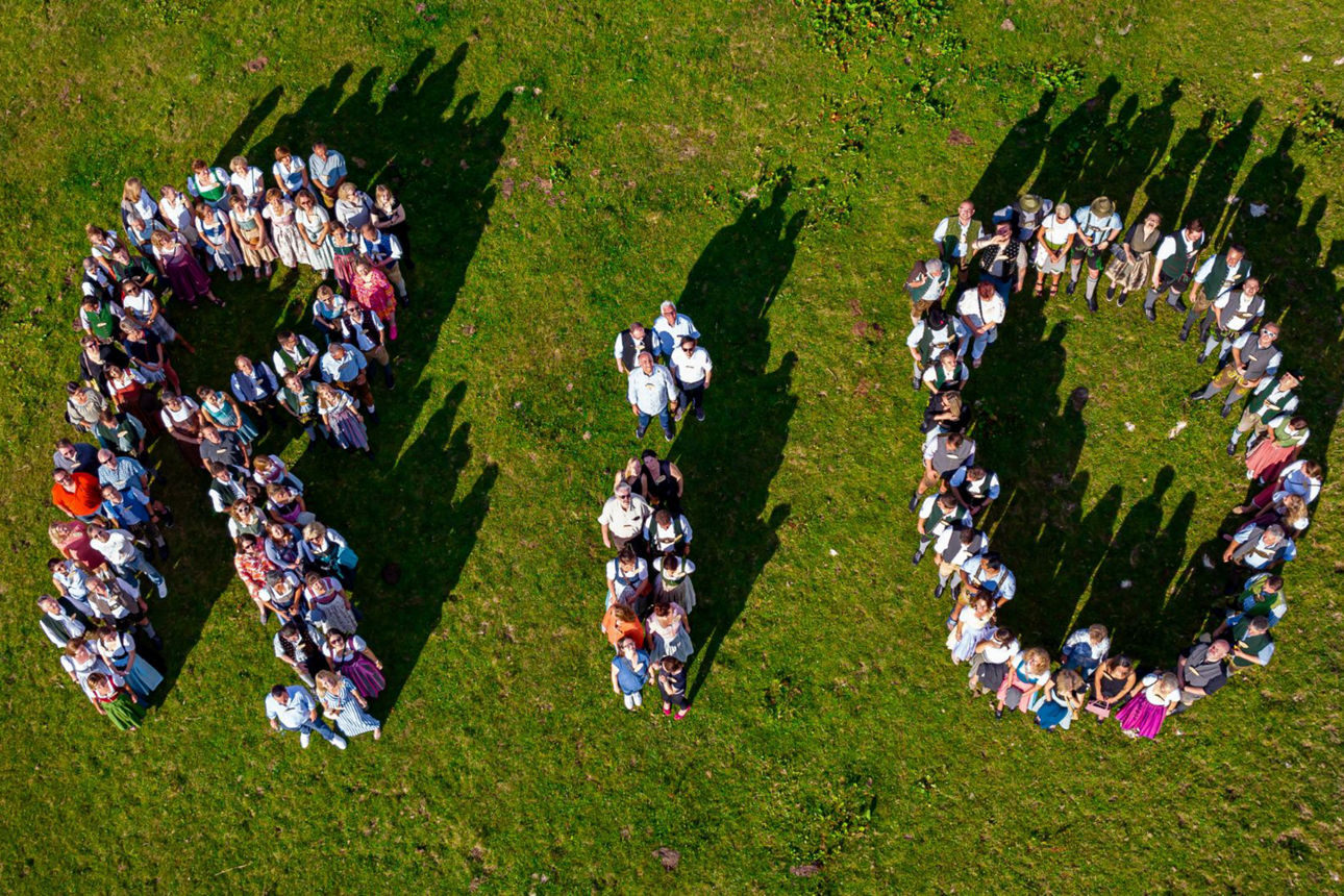 Eine Gruppe von Menschen in Trachtenkleidung bildet auf einer grünen Wiese die Buchstaben "R I O". Drei Kühe grasen in der Nähe. Die Aufnahme ist aus der Vogelperspektive gemacht.