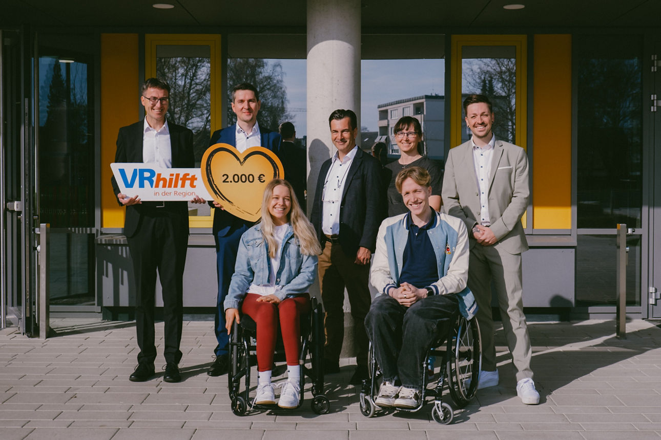 Zwei Personen sitzen lächelnd im Rollstuhl, dahinter mehrere Personen ebenfalls lächelnd mit einem Spendenscheck in der Hand.