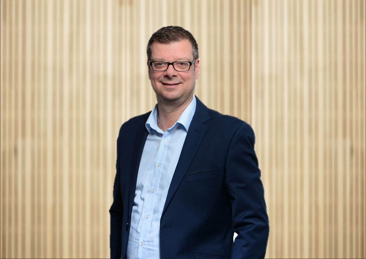 Mann mit blauem Hemd und dunkelm Sacko steht vor Holzwand