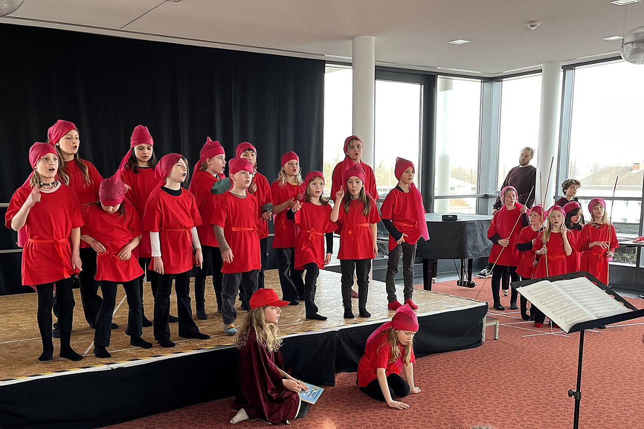 EIn Kinderchor, gekleidet mit roten Mützen, roten T-Shirts und schwarzen Hosen, steht auf einer Bühne, zwei Kinder sitzen davor, ein paar Kinder rechts neben der Bühne