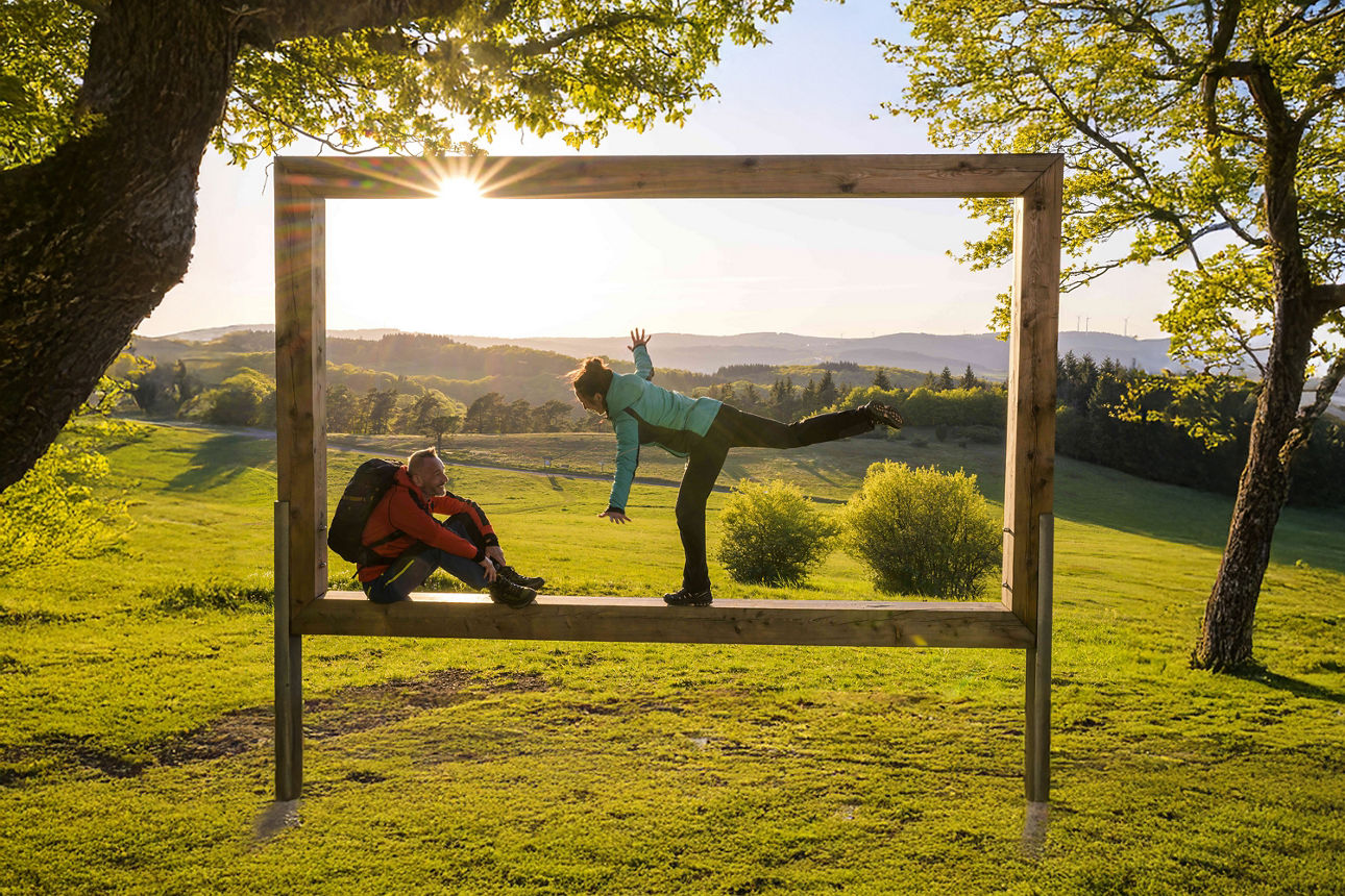 Zwei Wanderer posieren kreativ in einem großen Holzrahmen mit Aussicht auf eine malerische Hügellandschaft bei Sonnenuntergang. Der Rahmen ist Teil eines Erlebnispunkts entlang des Rothaarsteigs.