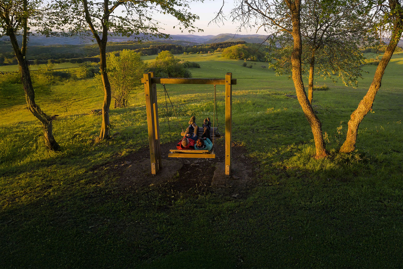 Zwei Personen liegen entspannt nebeneinander auf einer großen Holzschaukel und genießen die Aussicht auf die grünen Hügel und Wälder des Rothaarsteigs bei Sonnenuntergang