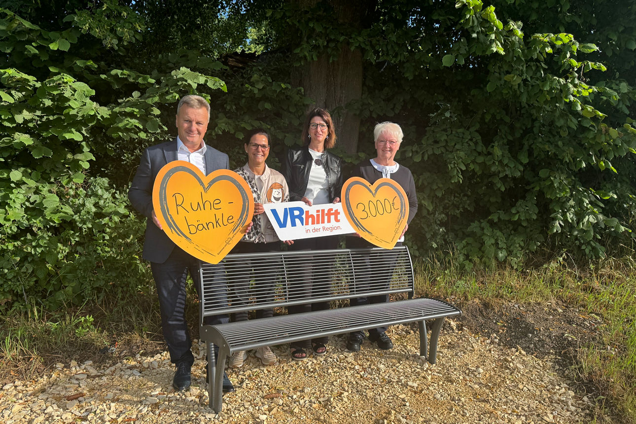 Drei Personen auf Parkbank mit Herzschildern und „VR hilft“-Schild im Grünen