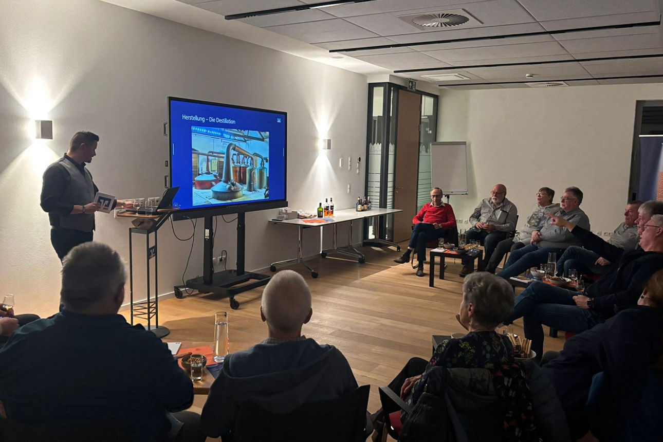 14 Personen sind in einem Raum. Ein Mann steht vor den anderen und erzählt etwas über die Herstellung von Rum in Form einer Präsentation..