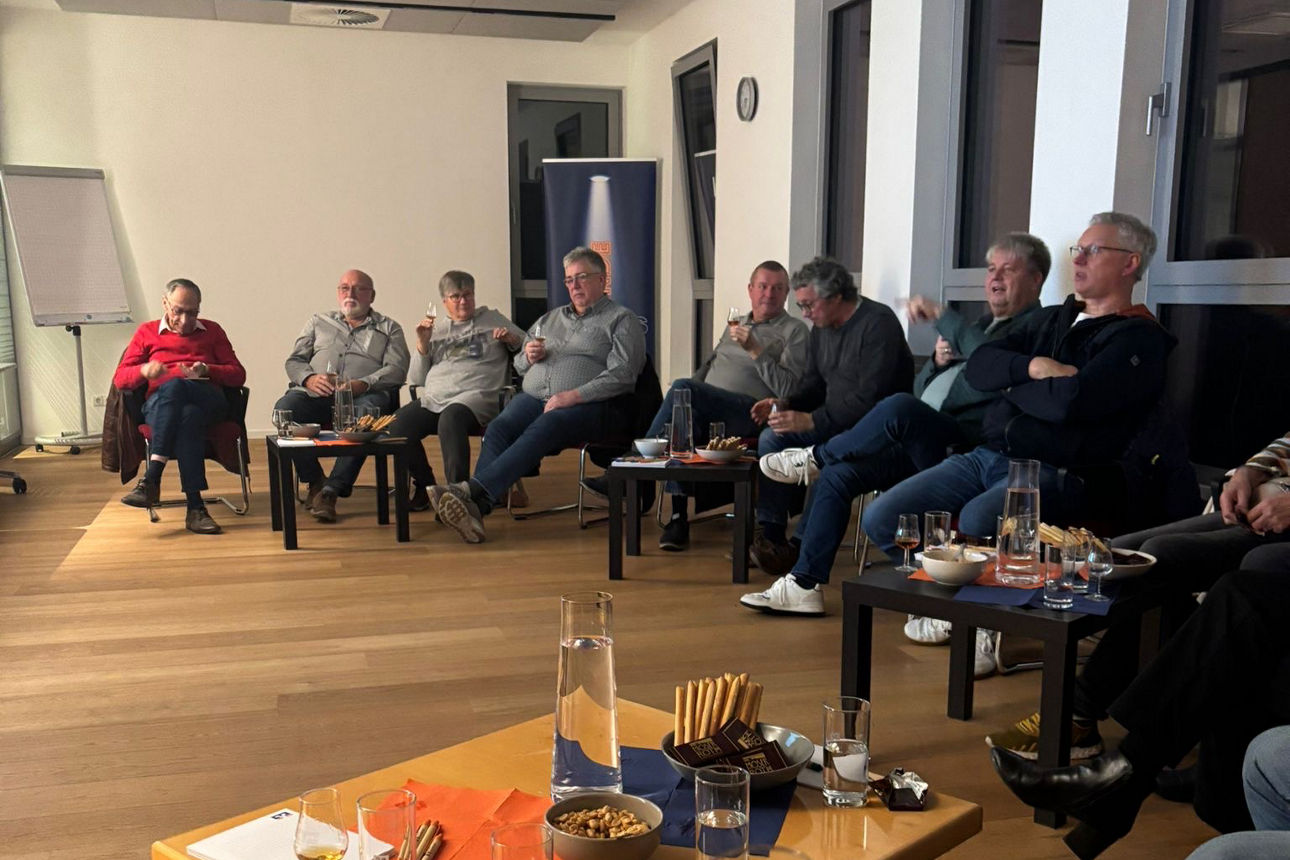 10 Personen sitzen in einem Raum mit Rum in der Hand. Vor ihnen sind gedeckte Tische mit Snacks.