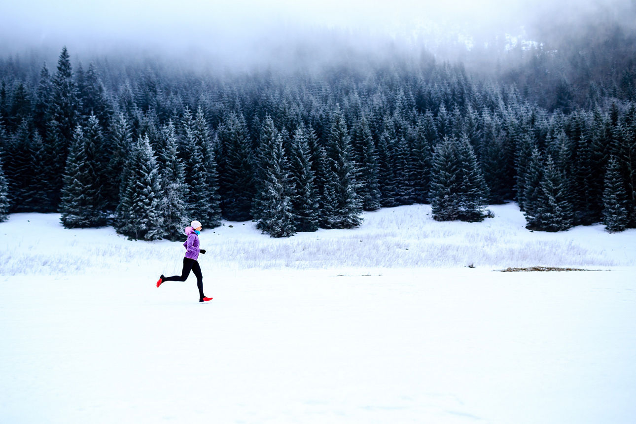Frau joggt im Schnee in bunter Kleidung vor Waldkulisse