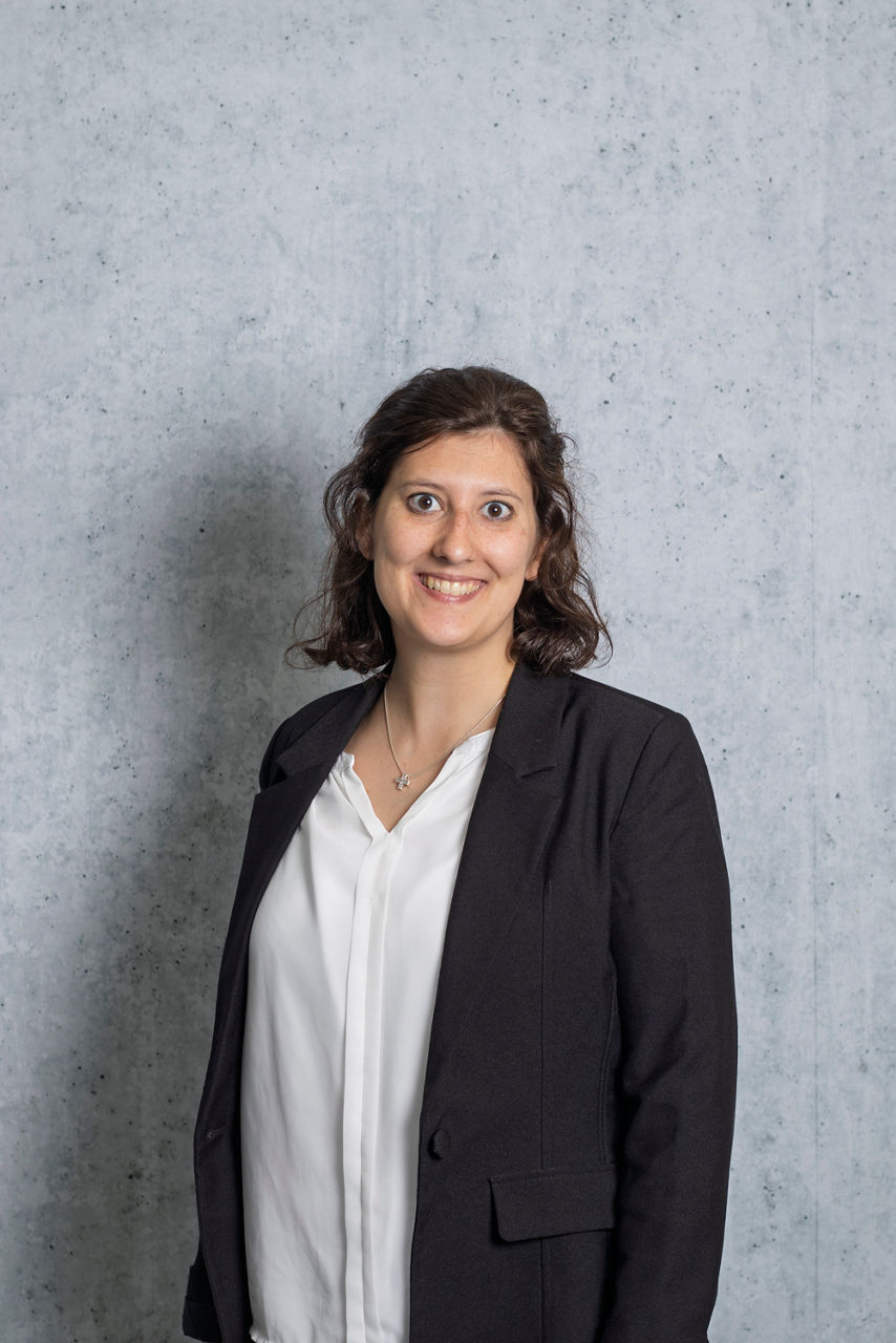 Junge Frau steht vor grauer Wand und sieht lächelnd in die Kamera