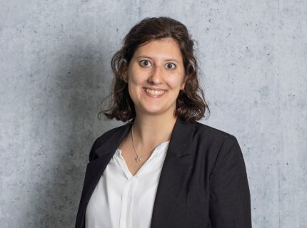 Profilbild einer jungen Frau, die lächelnd vor einer grauen Wand steht