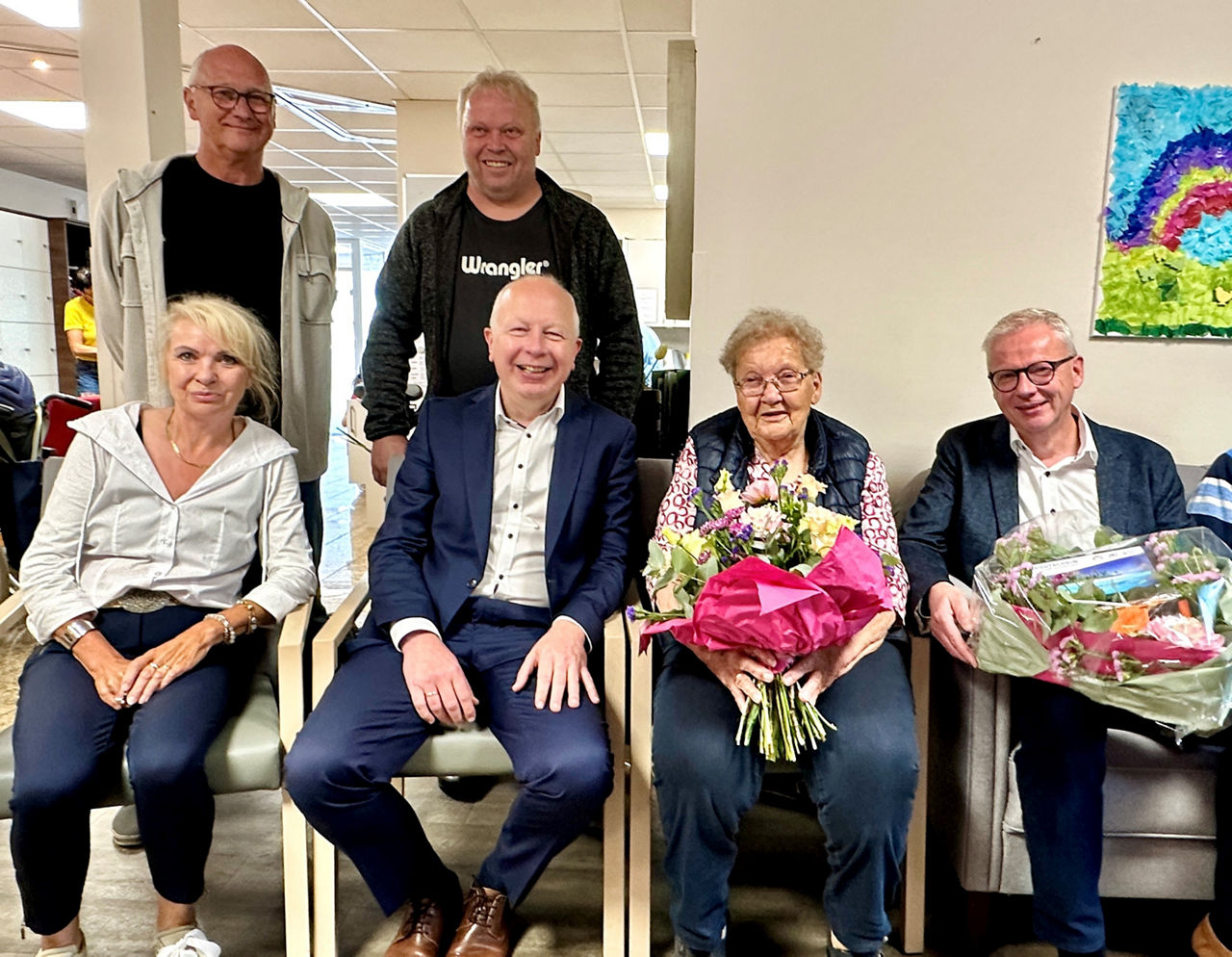 Mehrere Personen - 4 sitzen im Vordergrund - 2 stehen im Hintergrund - 2 Personen halten Geschenk und Blumen in der Hand