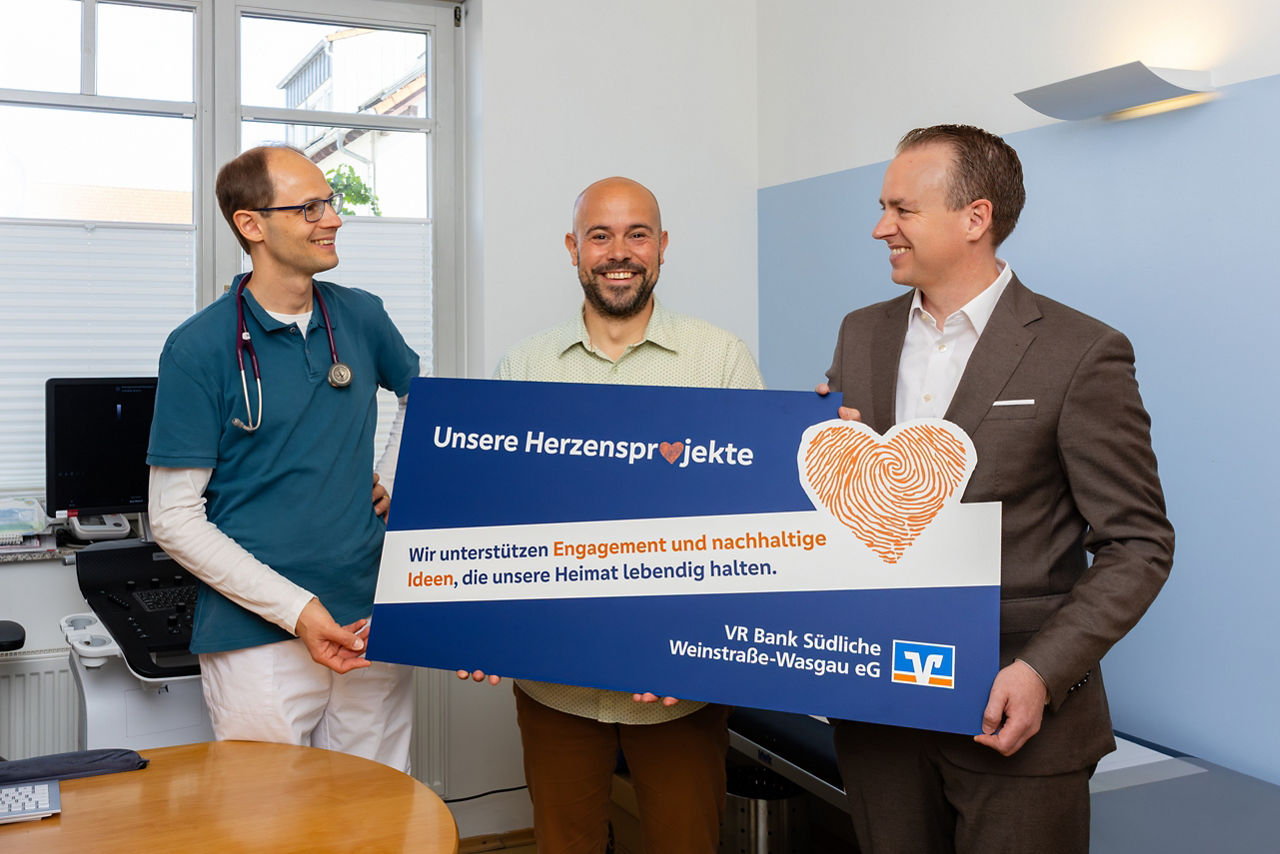 Drei Personen stehen in einem Raum. Sie halten gemeinsam einen großen Scheck. Im Hintergrund ist medizinische Ausstattung.
