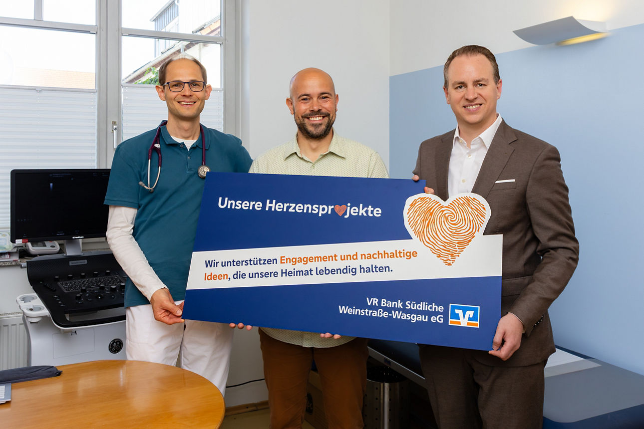 Drei Personen stehen in einem Raum. Sie zeigen zusammen einen großen Scheck. Im Hintergrund sind Geräte aus einer Arztpraxis.