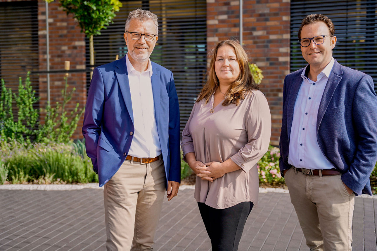 Drei Personen stehen vor einem Backsteingebäude und lächeln.