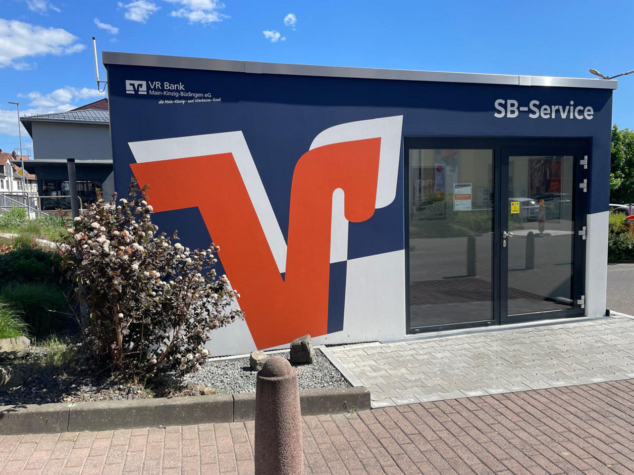 Das Bild zeigt den SB-Pavillon in Nidda. Er ist blau, hat eine große zweiflügelige Tür und auf ihm ist ein großes V in orange