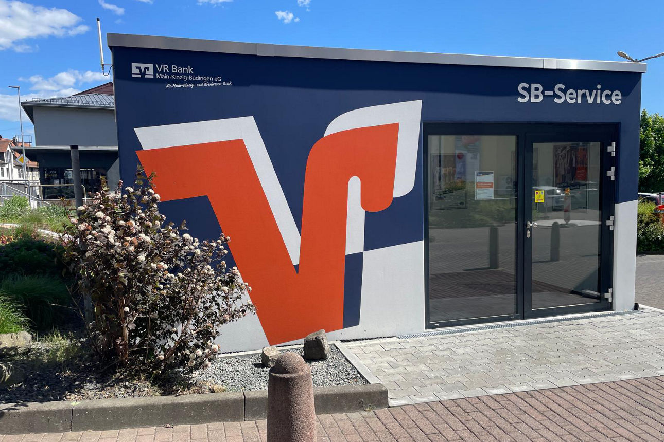 Das Bild zeigt den SB-Pavillon in Nidda. Er ist blau, hat eine große zweiflügelige Tür und auf ihm ist ein großes V in orange