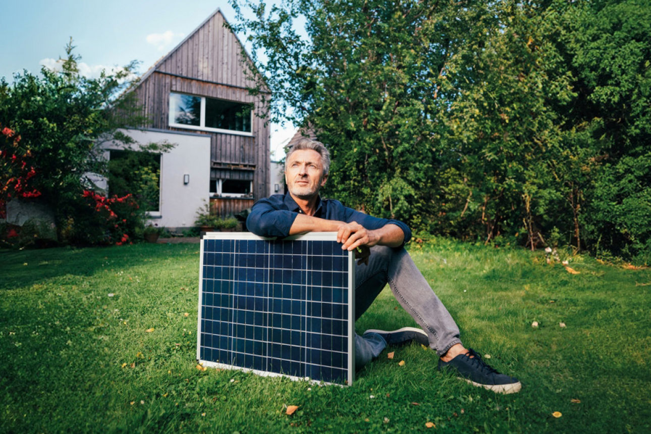 Ein Mann sitzt vor einem Haus mit einem Solarpanel im Arm - Modernisierungscheck